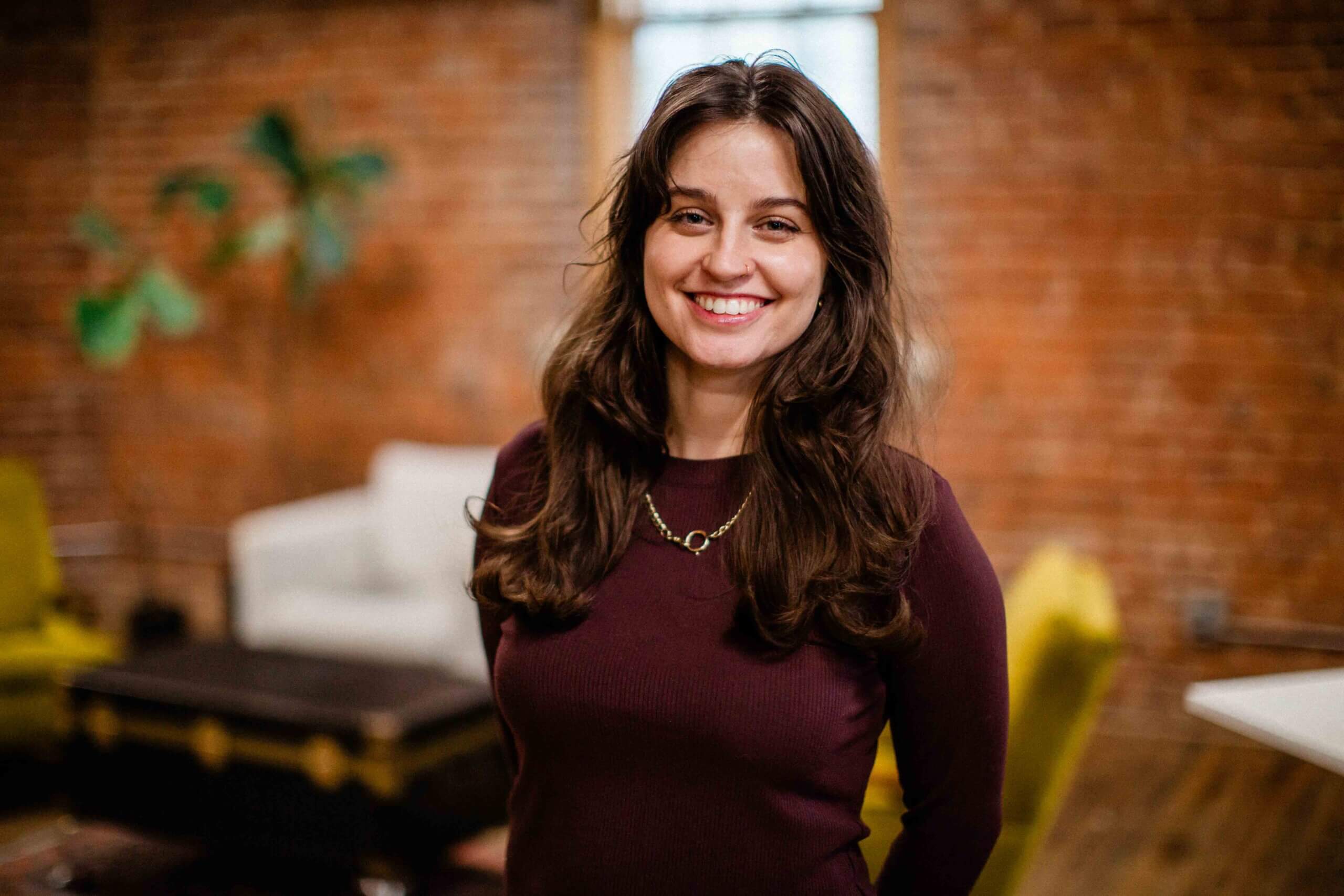 allison Smiling woman with long brown hair, wearing a maroon top, standing in a modern office space with exposed brick walls, reflecting her role as a detail-focused marketing strategist at Harris & Ward.