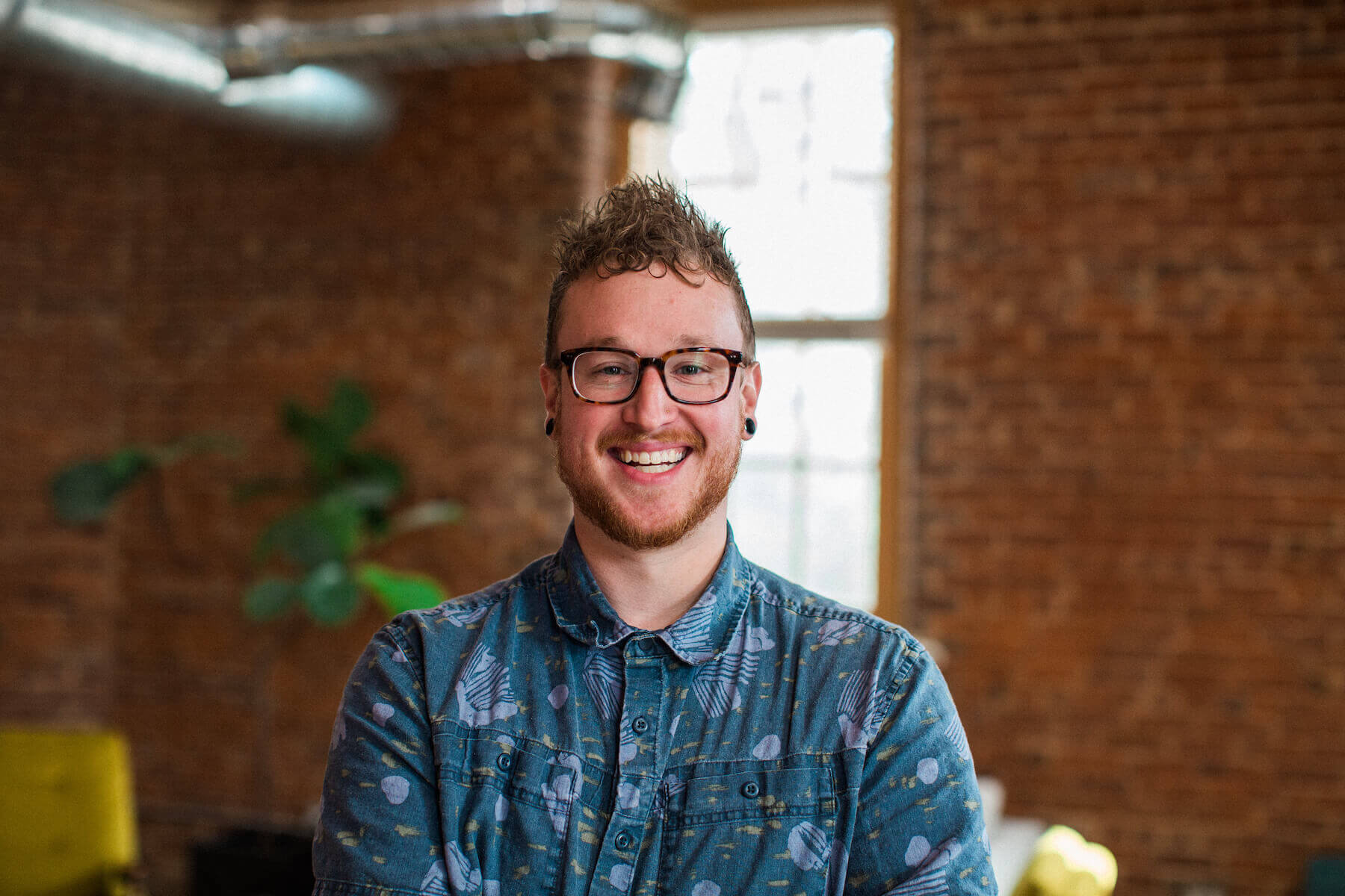 bradley Corey, partner at Harris & Ward, smiling in a casual button-up shirt, standing in a modern office with exposed brick and greenery in the background, representing the agency's commitment to quality and teamwork.