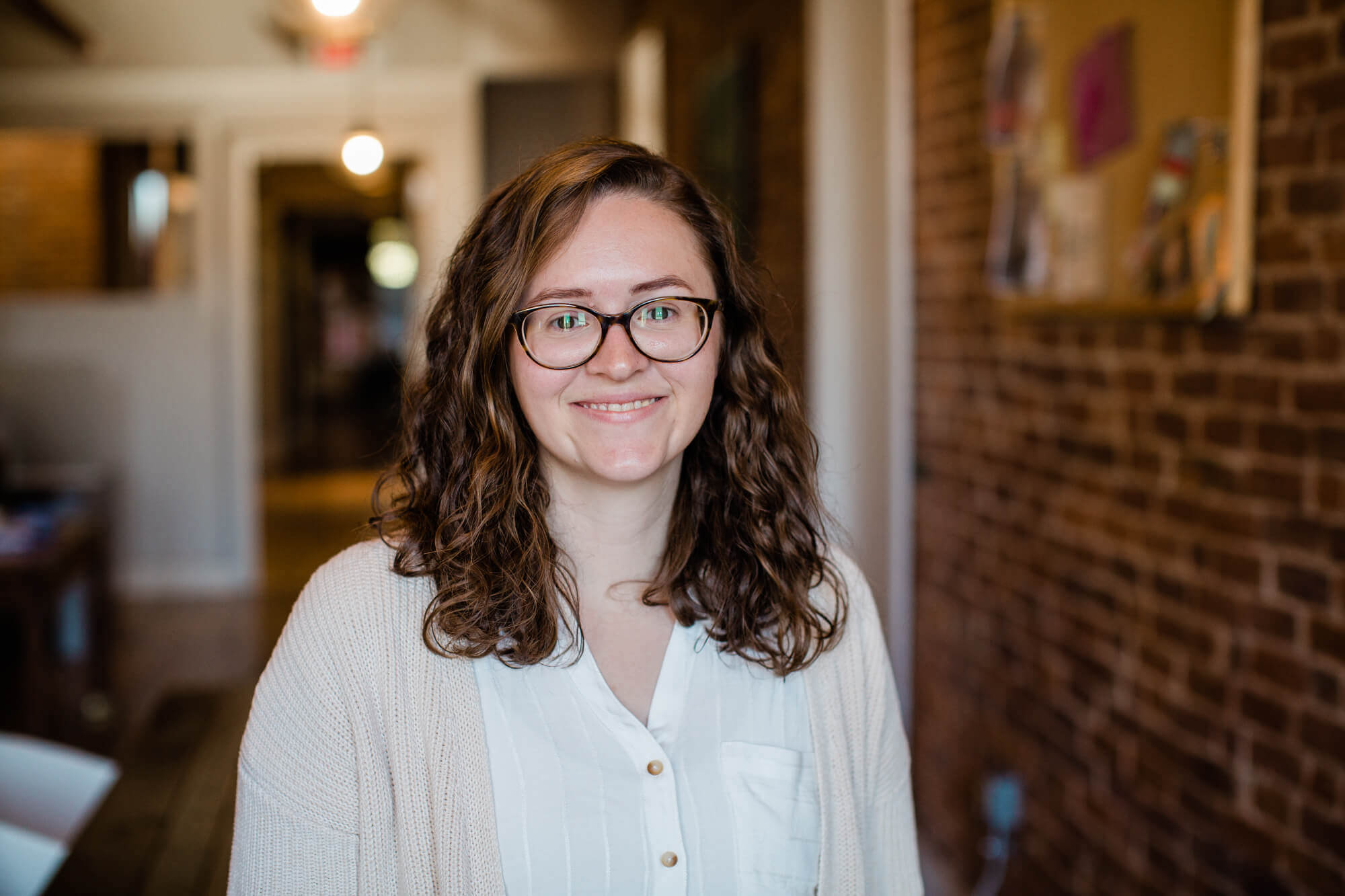 cailley Cailley Harris, Production Coordinator at Harris & Ward, smiling in a cozy office setting with brick wall background, showcasing her detail-oriented and creative approach to project coordination.