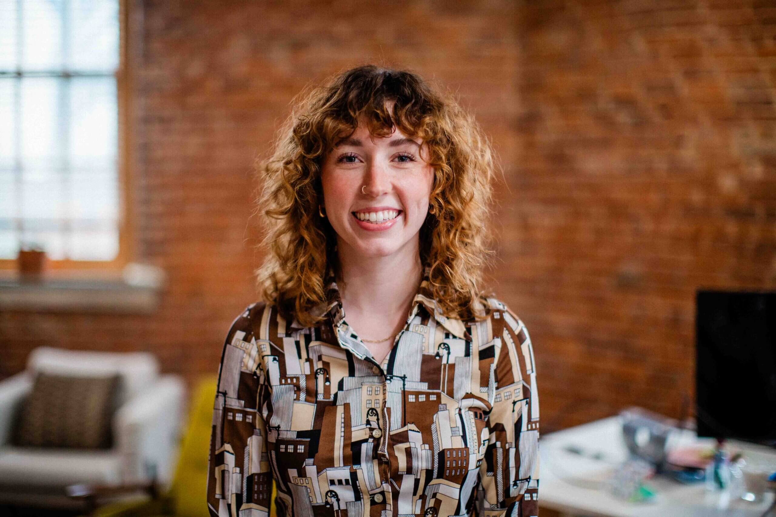 lindsey Lindsey Reister smiling in a vintage-style shirt, showcasing her project management skills and vibrant personality in a creative office setting at Harris & Ward.