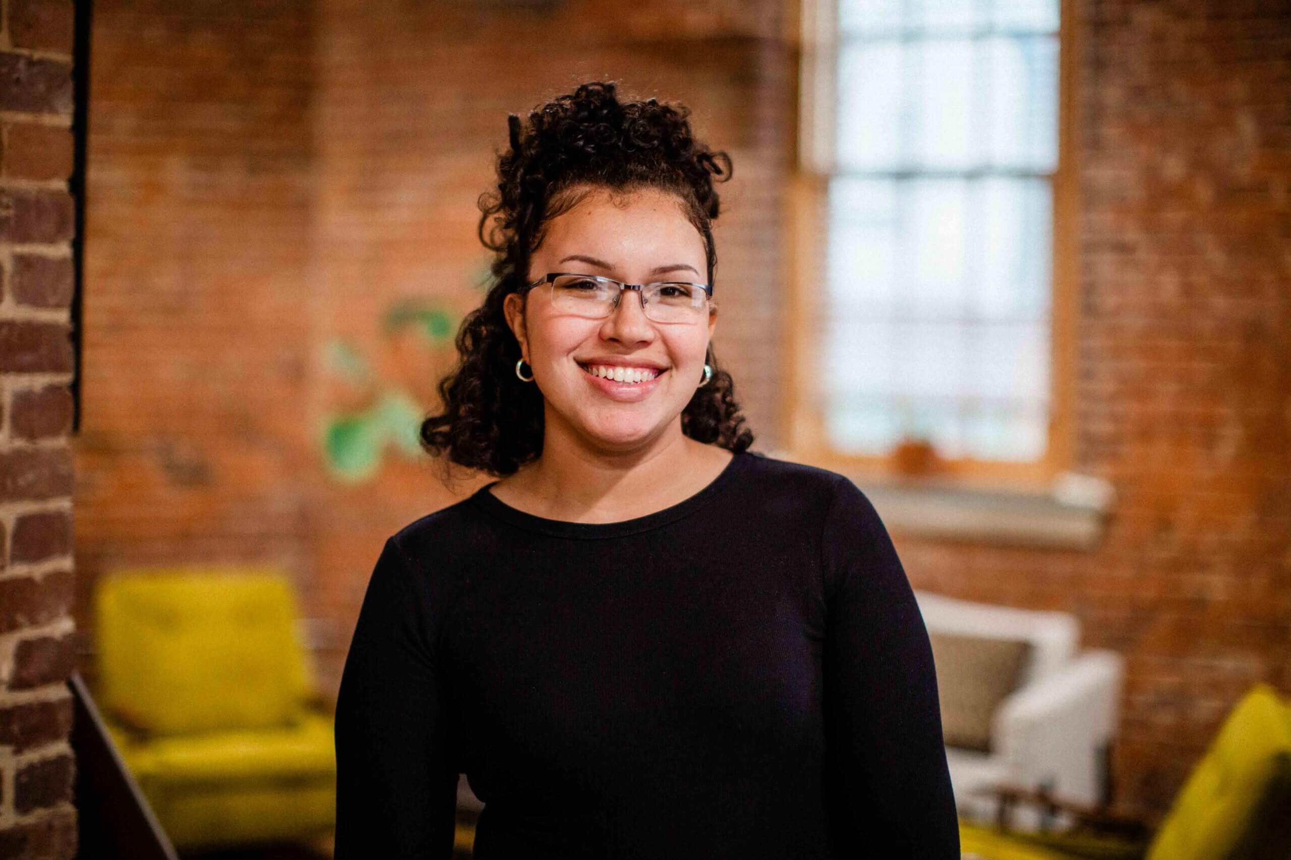 siera Siera Arena, Client Experience Specialist at Harris & Ward, smiling in a cozy, brick-walled office with vintage decor, showcasing her approachable personality and professional warmth.