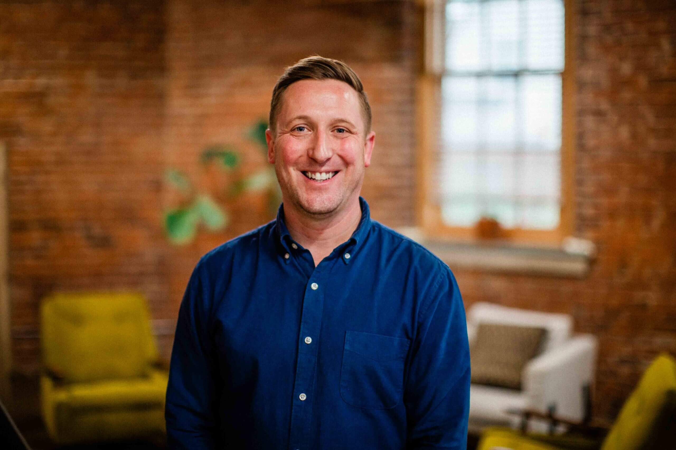 todd Todd Boak, partner at Harris & Ward, smiling in a blue shirt, set against a brick wall with green accents, embodying a creative marketing atmosphere in Lexington.